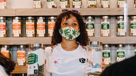 Photo of a women behind the counter and wearing a mask with pot leaves on it at the Kaleafa Cannabis business while helping serve a customer